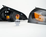 A small automotive amber bulb centered in the foreground, with two black headlight assemblies blurred in the background on a clean white studio surface.