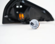 A small automotive light assembly component with a gray plastic housing and orange reflector, shown in sharp focus on a white surface with blurred black vehicle light housings in the background.