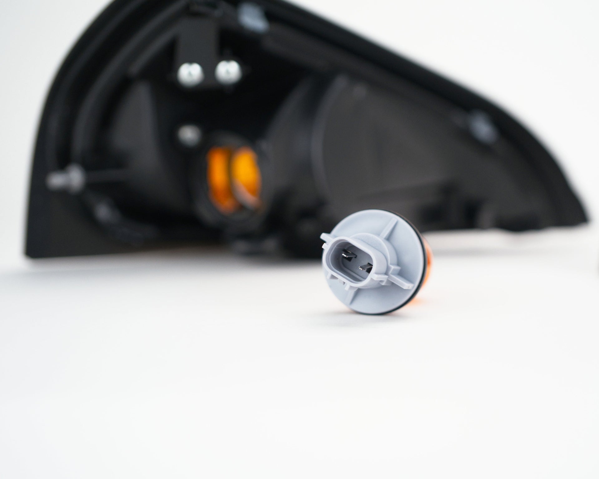 A small automotive light assembly component with a gray plastic housing and orange reflector, shown in sharp focus on a white surface with blurred black vehicle light housings in the background.