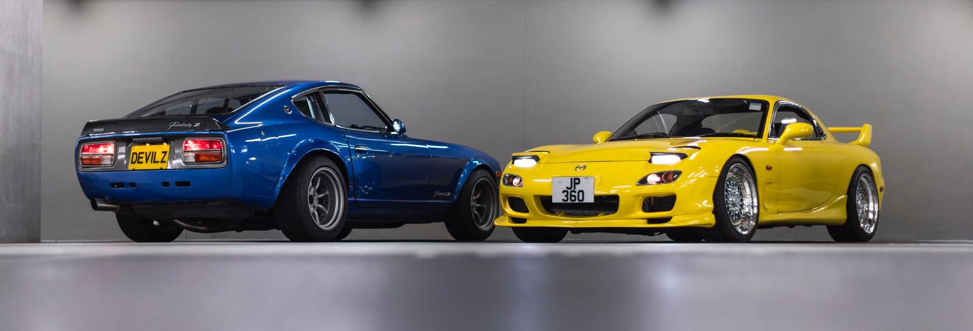Two sports cars parked side by side in a garage: a blue coupe on the left and a bright yellow coupe on the right, photographed from the rear and front angles respectively.